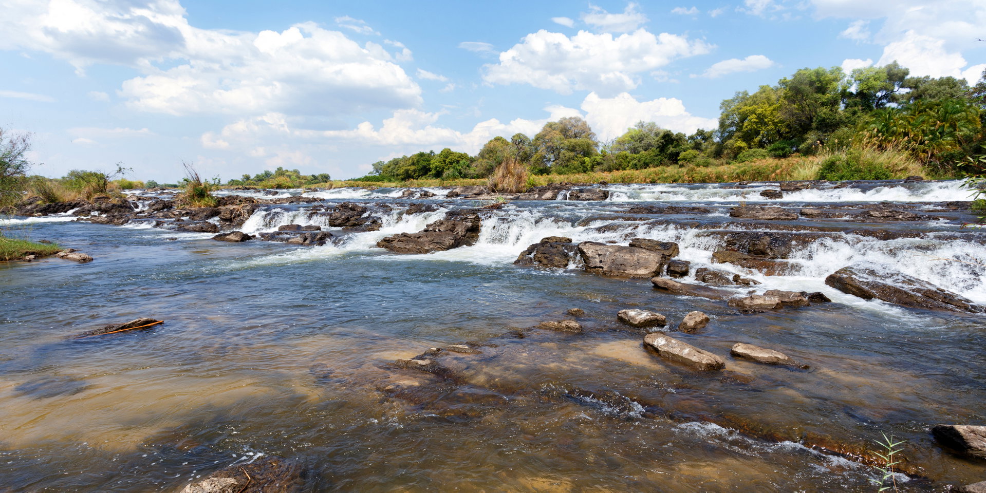 White water rushes over rocks between green riverbanks.