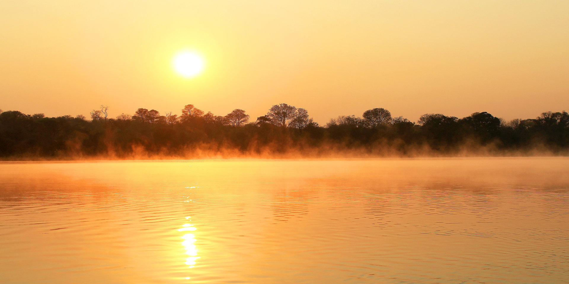 The sun sets in an orange sky over a calm river lined with trees.