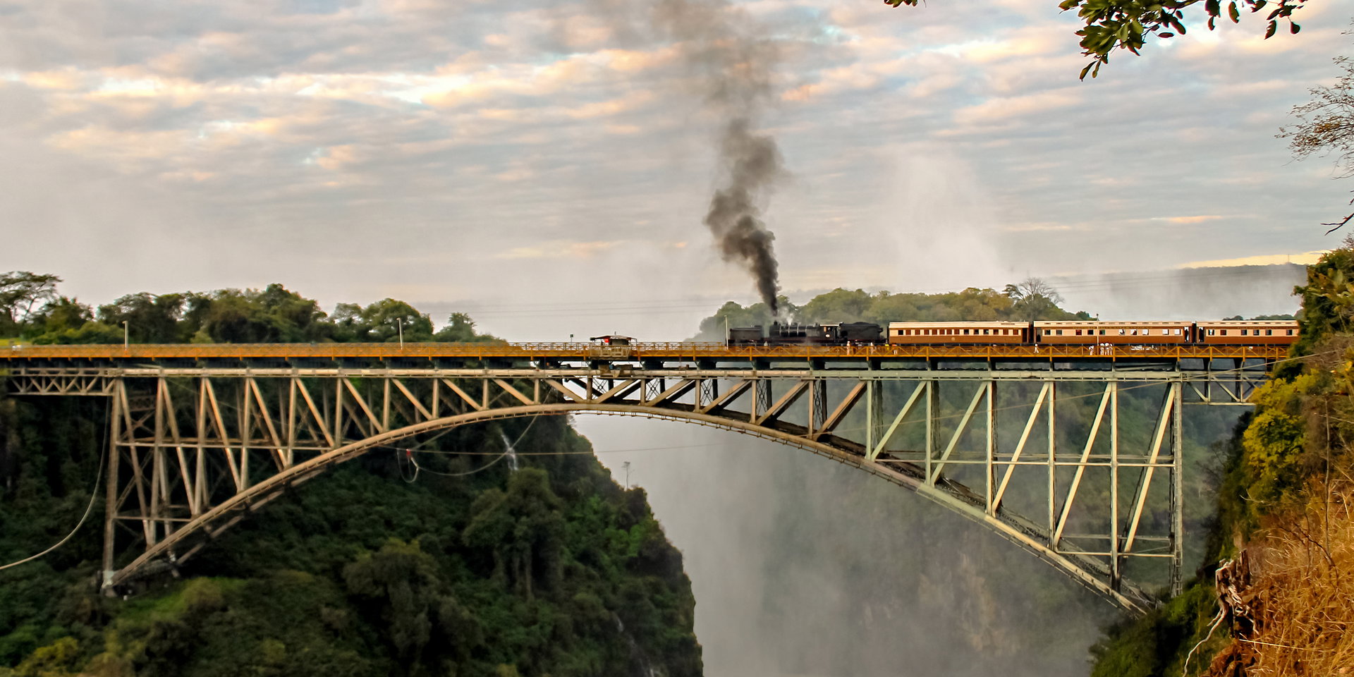 A steam train crosses the steel arch bridge above the misty gorge.